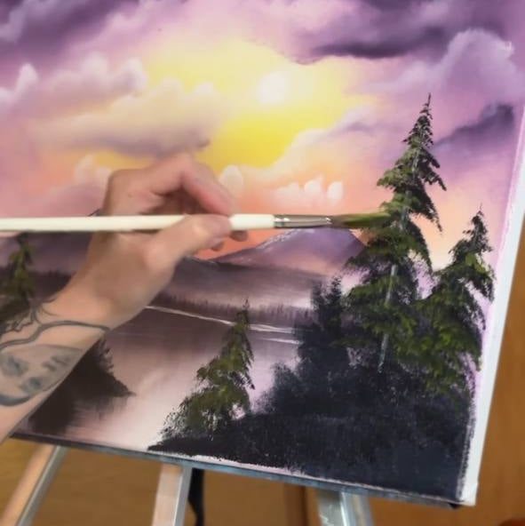 Person painting a landscape with trees and a sunset on an easel.